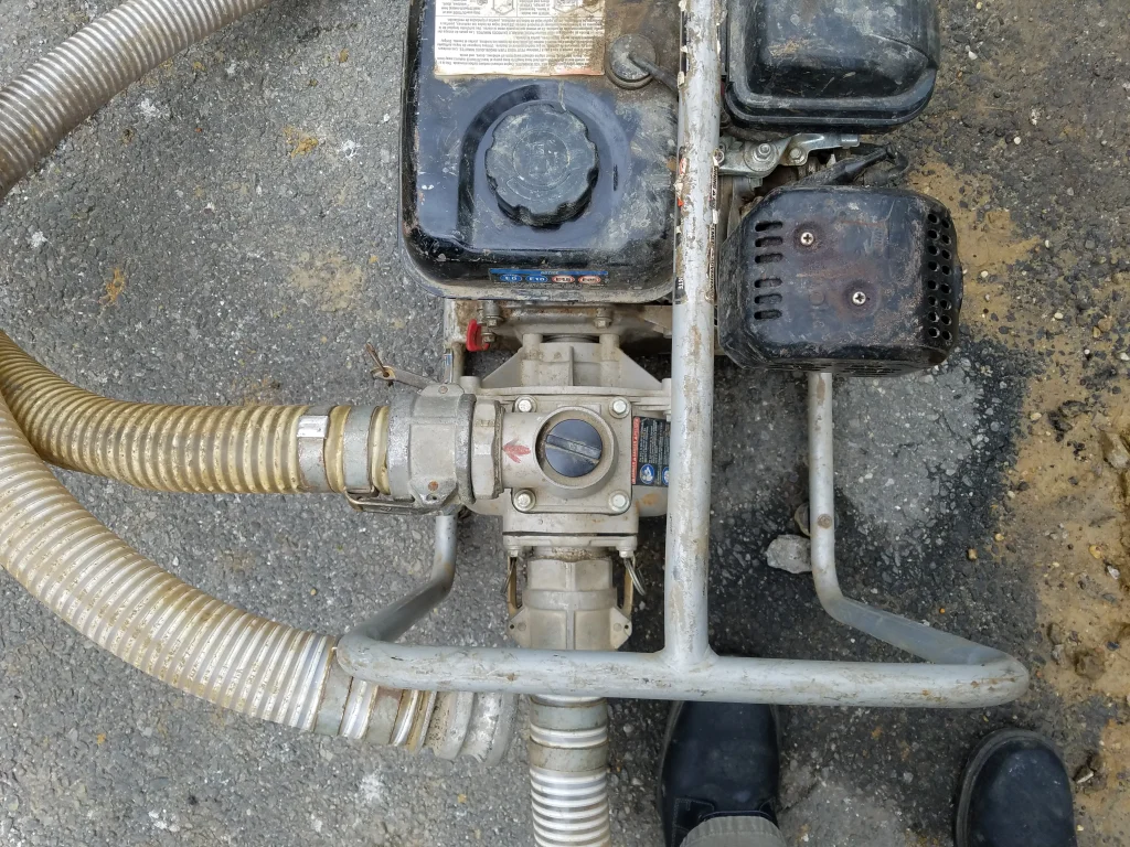 Top view of a portable water pump with hoses attached, placed on a rough asphalt surface. The pump is metallic and black, showing signs of wear and dirt. Two boots are partially visible at the bottom of the image.