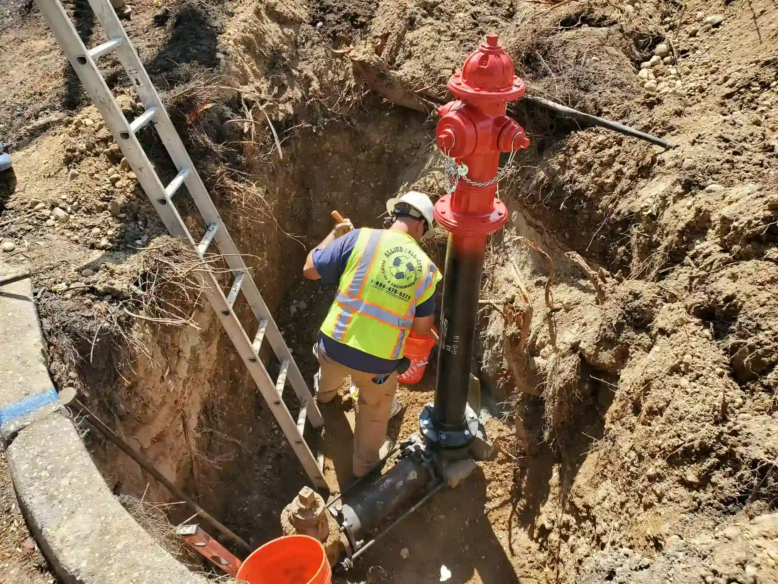 A worker wearing a safety vest and hard hat installs or maintains a red fire hydrant in a dug-out trench, with a ladder positioned nearby and construction tools and materials around.